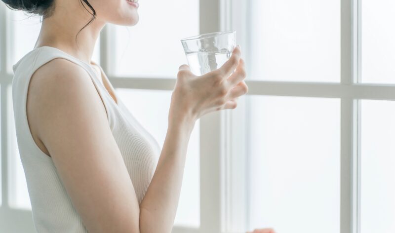 窓辺でグラスの水を手に、ほほ笑むノースリーブの女性