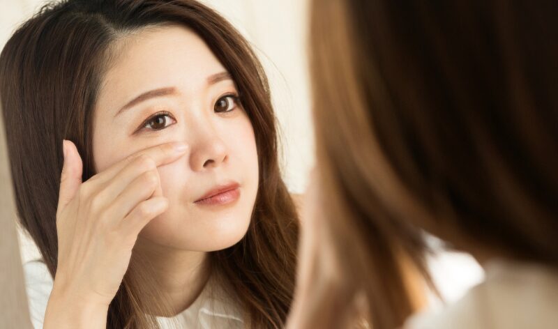 鏡を見ながら目の下にそっと手を添える女性。目元の変化に気づく瞬間。