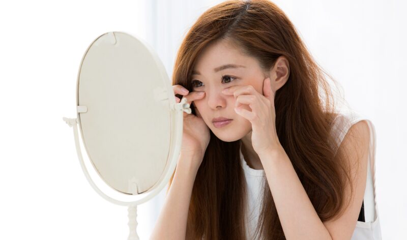 鏡の前で目元を指で強く引っ張る女性。過度な刺激による目元ケアのリスクを表現。
