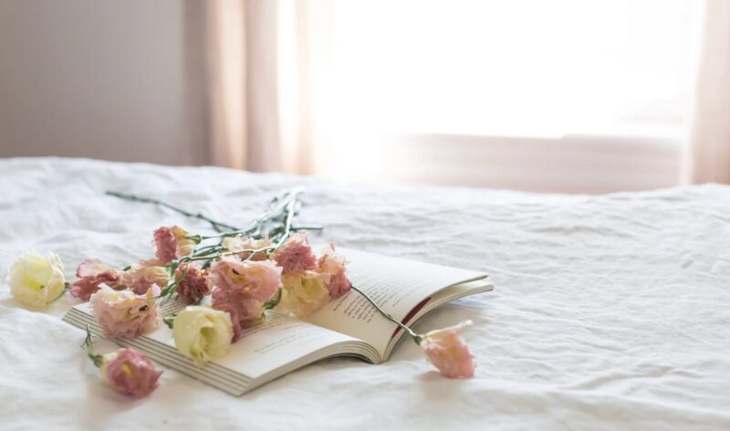 白いベッドの上に置かれた本とピンクの花。静かな朝のひとときを感じさせる、わたしに戻る時間の象徴。