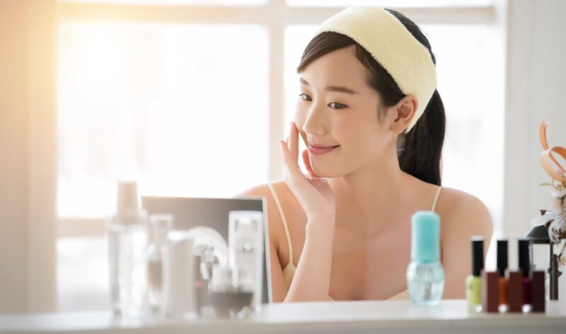 朝の光の中でスキンケアをする女性。鏡の前で微笑みながら肌を整えている様子。