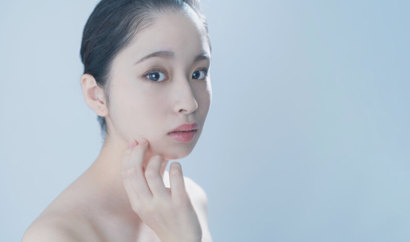 青い背景の中で素肌を見つめる女性。透明感のある肌を目指したシンプルケアのイメージ。